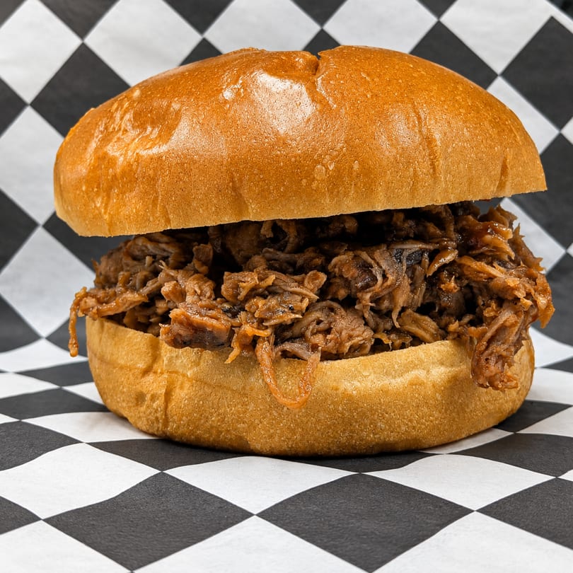 A pulled pork sandwich on a brioche bun, overflowing with meat, on black and white checkered paper.
