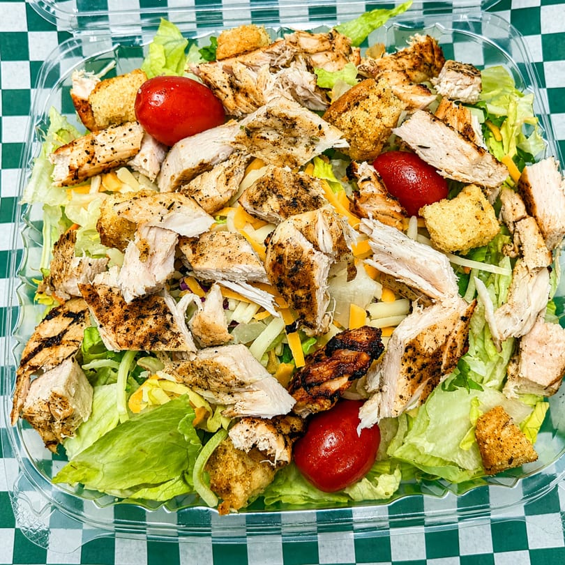 Grilled chicken salad with lettuce, shredded cheese, croutons, and cherry tomatoes in a plastic container.