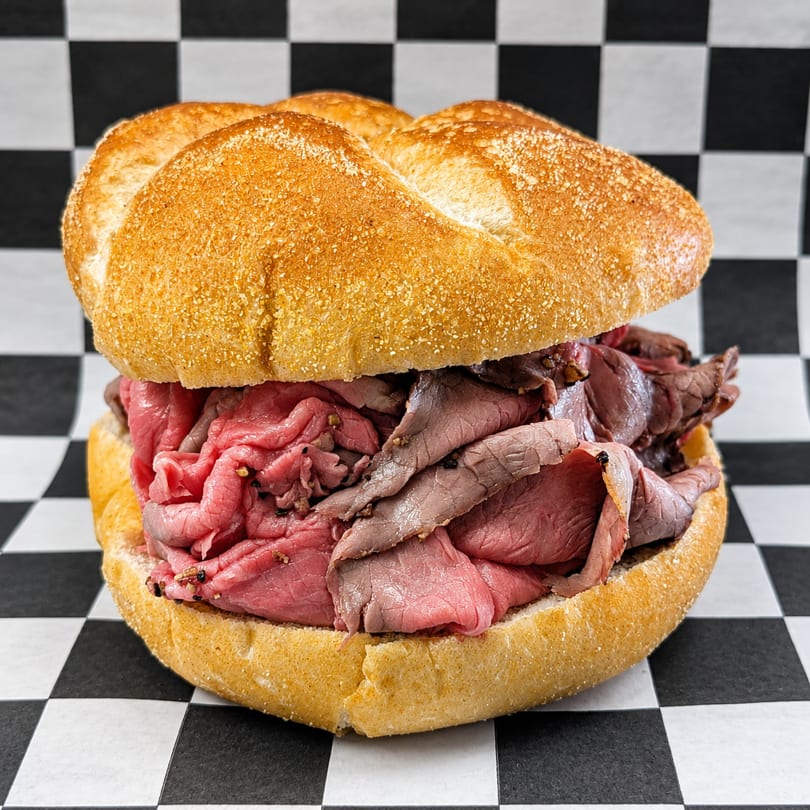 A large, fresh roast beef sandwich on a kaiser roll, against a black and white checkered background.
